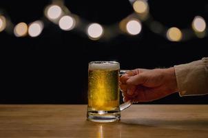 Hand, die ein Glas Bier auf einem Holztisch hält, auf dem Bokeh-Lichter mit dunklem Hintergrund sind. foto