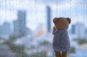 Teddybär weint allein am Fenster, wenn es mit verschwommenem Stadthintergrund regnet. foto