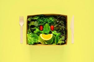 Salatset als lächelndes Gesicht in Einweg-, kompostierbare Lebensmittelbox aus Papier, Gabel und Löffel auf gelbem Hintergrund für das Konzept des Weltumwelttages. foto