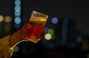 Hand mit einem Glas Bier, das zum Feiern und Partykonzept anstößt, isoliert auf dunklem Nachthintergrund mit bunten Bokeh-Lichtern der Stadt auf der Bar auf dem Dach. foto
