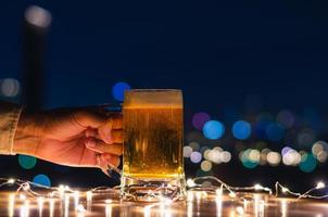 Hand, die ein Glas Bier auf Holztisch mit buntem Stadt-Bokeh-hellem Hintergrund hält. foto