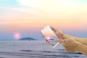 Hand, die ein mobiles Smartphone mit Strand- und Himmelshintergrund hält foto