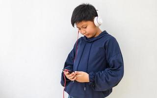 Süßer asiatischer Junge mit weißer Haut in einem Kapuzenpulli, der Musik auf einem Mobiltelefon und Kopfhörern isoliert über weißem Hintergrund hört. Porträt- und Kopierbereich foto