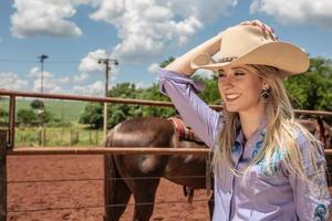 Schönes blondes Cowgirl mit Hut, das nahe dem Hintergrund der Pferderanch steht foto