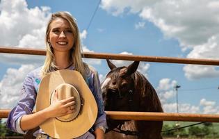 Schönes blondes Cowgirl mit Hut, das nahe dem Hintergrund der Pferderanch steht foto