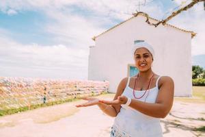 Fröhliche Brasilianerin in traditioneller bahianischer Tracht, die etwas auf ihrer Handfläche zeigt, blickt in die Kamera, mit dem historischen Zentrum von Porto Seguro im Hintergrund foto