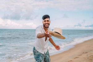 lateinamerikanischer mann lächelt und wirft seinen hut, der an einem schönen sommertag auf die kamera am strand schaut foto