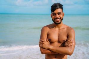 athletischer junger lateinamerikanischer Mann am Strand mit verschränkten Armen. lächelnder Mann, der in die Kamera schaut