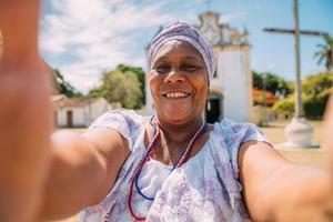 Fröhliche Brasilianerin afrikanischer Abstammung in traditioneller bahianischer Kleidung, die vor der Kirche ein Selfie macht. Fokus auf Gesicht