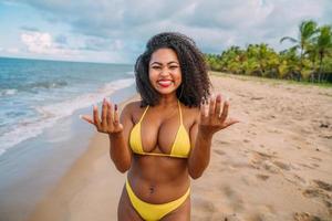 schöne lateinamerikanische Frau im Bikini am Strand. junge frau, die an einem sonnigen tag sommerferien genießt, lächelt, zum strandbesuch einlädt und in die kamera schaut foto