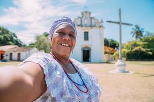 Fröhliche Brasilianerin afrikanischer Abstammung in traditioneller bahianischer Kleidung, die vor der Kirche ein Selfie macht. Fokus auf Gesicht