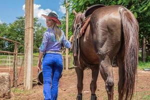 Schönes blondes Cowgirl mit Hut, das rückwärts in der Nähe des Hintergrunds der Pferderanch steht foto