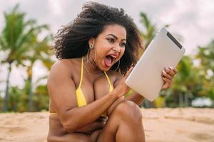 Frau, die im Sommerurlaub einen Videoanruf tätigt. lateinamerikanische frau, die mit einem tablet auf dem sand sitzt foto