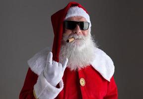 weihnachtsmann mit sonnenbrille, der das rocker-handzeichen zeigt und eine zigarre auf dunklem hintergrund raucht foto