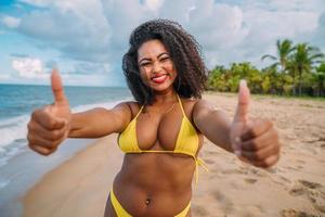 schöne lateinamerikanische Frau im Bikini am Strand. junge frau, die ihren sommerurlaub an einem sonnigen tag genießt, lächelt, mit daumen hoch und in die kamera schaut foto