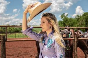 Schönes blondes Cowgirl mit Hut, das nahe dem Hintergrund der Pferderanch steht foto