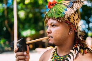 Indianerhäuptling des Pataxo-Stammes, der Pfeife raucht. brasilianischer Indianer mit Federschmuck und Halskette foto