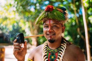 Indianerhäuptling des Pataxo-Stammes, der Pfeife raucht. Brasilianischer Indianer mit Federkopfschmuck und Halskette mit Blick auf die Kamera foto