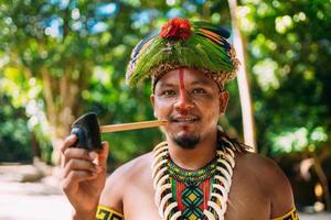 Indianerhäuptling des Pataxo-Stammes, der Pfeife raucht. Brasilianischer Indianer mit Federkopfschmuck und Halskette mit Blick auf die Kamera foto