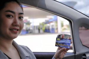 Fröhliche schöne asiatische Frau, die in ihrem Auto sitzt und Kreditkartenzahlung an einer Tankstelle zeigt foto