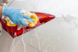 Frauenhand, die blaue Handschuhe mit gelbem Schwamm trägt, der modernes Auto des Rücklichts wäscht oder Auto reinigt. Autowaschkonzept foto