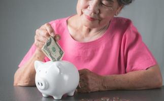 alte Seniorin, die Dollarnoten in ein weißes Sparschwein auf den Tisch legt. foto