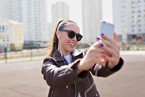 Attraktive junge Frau mit schwarzer Sonnenbrille und Lederjacke, die sich im Sonnenlicht auf dem Hintergrund der Stadt mit wunderbarem Lächeln, wahren Emotionen, selbst macht foto