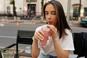 verführerische junge Frau, die in die Kamera zwinkert und lächelt, während sie Sommer-Smoothie trinkt und auf der Terrasse sitzt, Nahaufnahme. Auffallend sind die nackten und dünnen Rundungen der Schlüsselbeine. foto