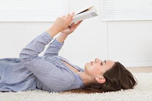 Frau, die Buch liest, während sie zu Hause auf Teppich liegt