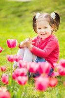 fröhliches kleines Mädchen, das im Gras sitzt und Tulpen betrachtet foto