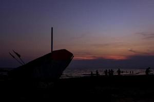 Silhouette des Bootes auf der linken Seite und diejenigen, die sich am Strand und dem magentafarbenen Dämmerungshimmel entspannen. foto