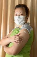 asiatische frau, die eine maske trägt und nach der impfung gegen covid-19, coronavirus-impfkonzept, pflaster auf ihrem arm zeigt. foto