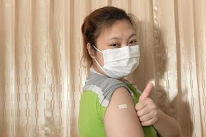 asiatische frau, die eine maske trägt und nach der impfung gegen covid-19, coronavirus-impfkonzept, pflaster auf ihrem arm zeigt. foto