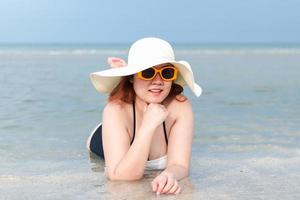 eine rundliche weiße frau in badeanzug, weißem hut und gelber sonnenbrille liegt lächelnd am strand. foto