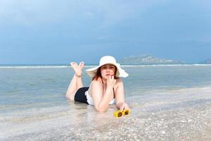 eine rundliche weiße frau in badeanzug oder badeanzug, weißem hut und gelber sonnenbrille liegt lächelnd am strand. foto