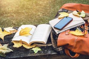 Der Rucksack und das Buch ruhen auf einem antiken Holztisch, Notizbüchern, Telefonen und der Natur der Ahornblätter von oben.