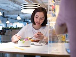 asiatische frau, die getrennt im restaurant sitzt und essen mit einer tischschild-kunststofftrennwand isst, um eine infektion vor coronavirus covid-19 zu schützen foto