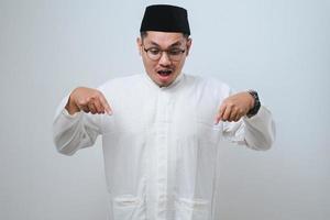 asiatischer muslimischer mann, der sich schockiert, mit offenem mund und erstaunt fühlt, ungläubig und überrascht vor weißem hintergrund nach unten schaut und zeigt foto