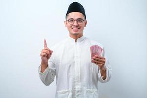 asiatischer muslimischer mann, der rupiah-papiergeld hält, während er mit dem finger nach oben zeigt und lächelt foto