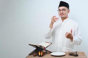 asiatischer muslimischer Mann lächelt und Daumen hoch, während er ein Glas Wasser zum Fastenbrechen trinkt foto