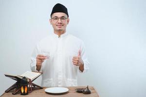 asiatischer muslimischer Mann lächelt und Daumen hoch, während er ein Glas Wasser zum Fastenbrechen trinkt foto