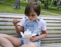 Ein junges Mädchen, das an einem Sommertag im Park spazieren geht, isst mit Nudeln Wok zu Mittag foto