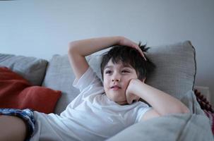 Porträt kleiner Junge mit neugierigem Gesicht, der auf dem Sofa sitzt und Zeichentrickfilme anschaut, Schulkind, das allein auf der Couch sitzt und sich nach der Schule im Wohnzimmer entspannt. foto