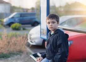 Kind, das an der Bushaltestelle sitzt und auf den Schulbus wartet. Porträt eines jungen Jungen, der mit denkendem Gesicht in die Kamera schaut, Schulkind, das an einem sonnigen Tag im Frühling oder Sommer an der Bushaltestelle im Freien sitzt foto