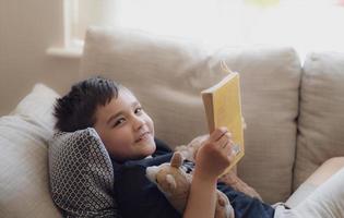porträtschulkind, das ein buch für hausaufgaben liest, kleiner junge, der auf sofa liegt, liest gerne die geschichte im wohnzimmer, kind entspannt sich zu hause an einem sonnigen frühlingstag, hausunterricht oder bildungskonzept foto