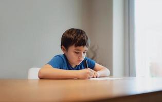 Kind, das auf dem Tisch sitzt und Hausaufgaben macht, Kind, Junge, der schwarzen Stift hält und auf weißem Papier schreibt, kleiner Junge, der zu Hause englische Wörter übt. Grundschule und Hausunterricht, Fernunterrichtskonzept foto