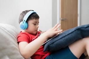 glücklicher kleiner Junge mit Kopfhörern, um mit Freunden im Internet zu spielen, Kind sitzt auf dem Sofa und liest oder schaut sich Cartoons auf dem Tablet an, Schulkind macht Hausaufgaben online zu Hause. foto