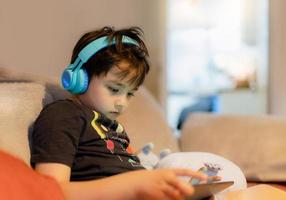 Fröhlicher kleiner Junge mit Kopfhörern zum Online-Spielen im Internet mit Freunden, Kind, das auf dem Sofa sitzt und liest oder Zeichentrickfilme auf Tablett-Kindern sieht, die sich morgens am Wochenende zu Hause entspannen foto