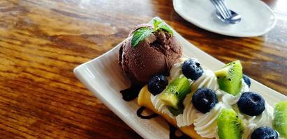 schokoladeneis mit geschnittener frischer kiwi, beere oder himbeere und schlagsahne in weißer platte oder teller auf holztisch mit kopierraum im café. dessert essen mit frischem obstkonzept foto