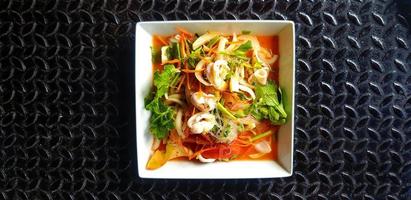thailändischer würziger Meeresfrüchtesalat mit Glasnudeln, Tintenfisch, Garnelen, geschnittenen Tomaten, Zwiebeln, Karotten und Sellerie auf weißem Teller oder Teller. asiatisches und berühmtes essen auf schwarzem edelstahlhintergrund oder tapete. foto